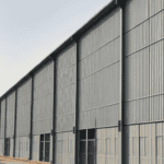 Steel salt barn exterior with corrugated metal panels and large glass windows, situated in an industrial area with a clear sky.