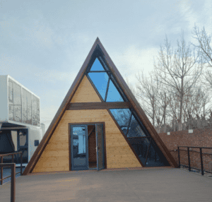 Ein modernes dreieckiges Hausdesign mit großen Glasplatten und Holzwänden, das eine stilvolle und einzigartige Ästhetik mit einem sauberen Terrassenbereich davor bietet.