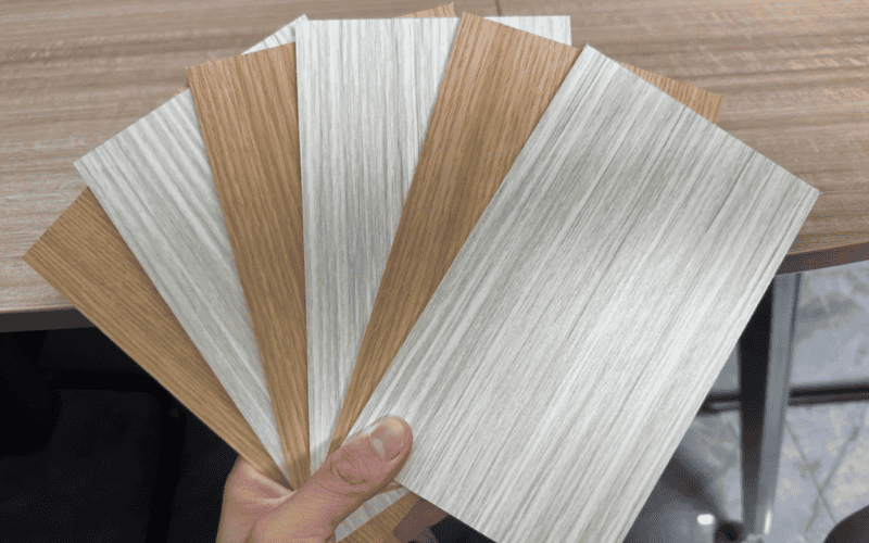 A hand holding multiple wood-patterned PVC panels, arranged like a fan, showcasing different shades and textures, placed on a wooden table.