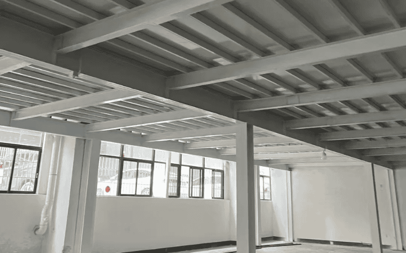 An interior view of a building's steel floor system, featuring grey steel beams and girders supporting the structure, with windows allowing natural light.