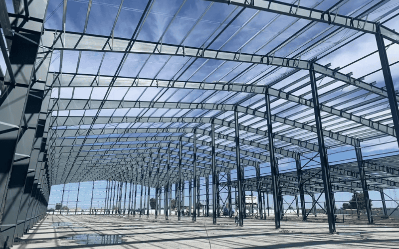 A spacious steel warehouse frame with a high ceiling, showcasing evenly spaced steel columns and beams under a translucent roof.