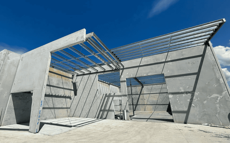 A modern industrial building under construction, with concrete walls and an exposed steel roof frame, set against a bright blue sky.