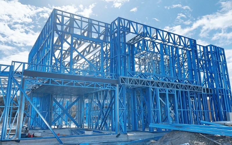 A two-story building under construction using blue steel framing, designed for a residential or commercial purpose, with a clear sky in the background.