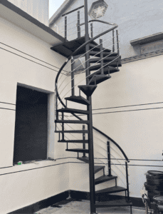 Outdoor spiral steel staircase with black railings, leading to an upper level of a building.