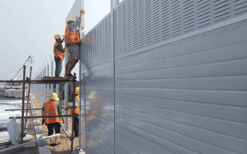 Workers installing a metal noise barrier with reflective and acoustic panels on a construction site.