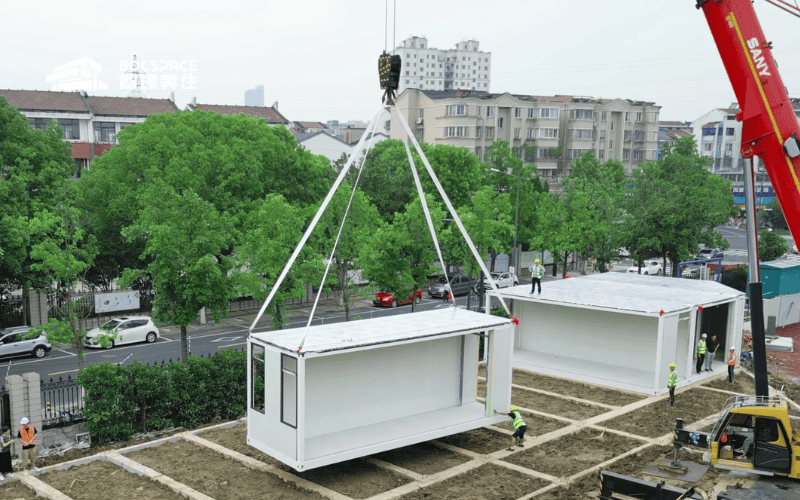 Workers using a crane to lift and position prefabricated modular steel buildings on-site, with surrounding green trees and city buildings in the background.