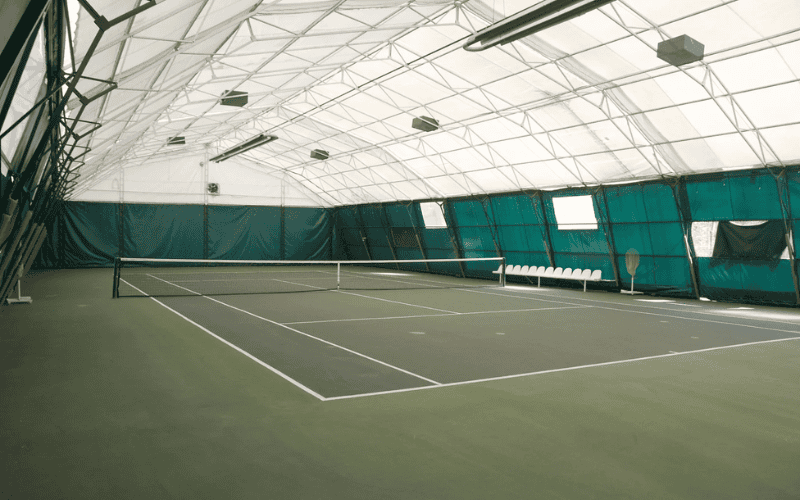 An indoor tennis court inside a fabric structure, with green walls and a net set up for a match.