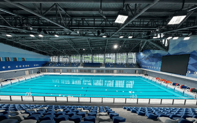 An indoor swimming pool with clear blue water, surrounded by seating and large windows in a modern facility.