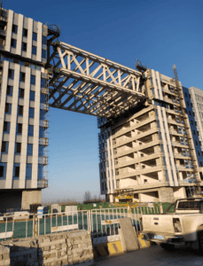 Construction of a steel truss bridge connecting two high-rise buildings, with scaffolding and clear blue skies in the background.