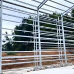 Partial view of a steel frame for a poultry building under construction, set against a backdrop of trees and earth mounds.