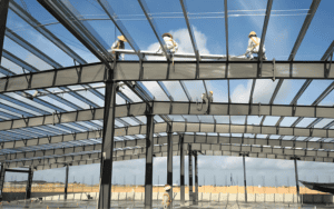 Workers in safety gear assembling a steel frame roof structure at a construction site under a clear blue sky.