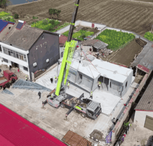 Eine Luftaufnahme eines Fertighauses, das mit einem Kran zusammengebaut wird. Arbeiter positionieren die vorgefertigten Module vor Ort, im Hintergrund sind Felder zu sehen.