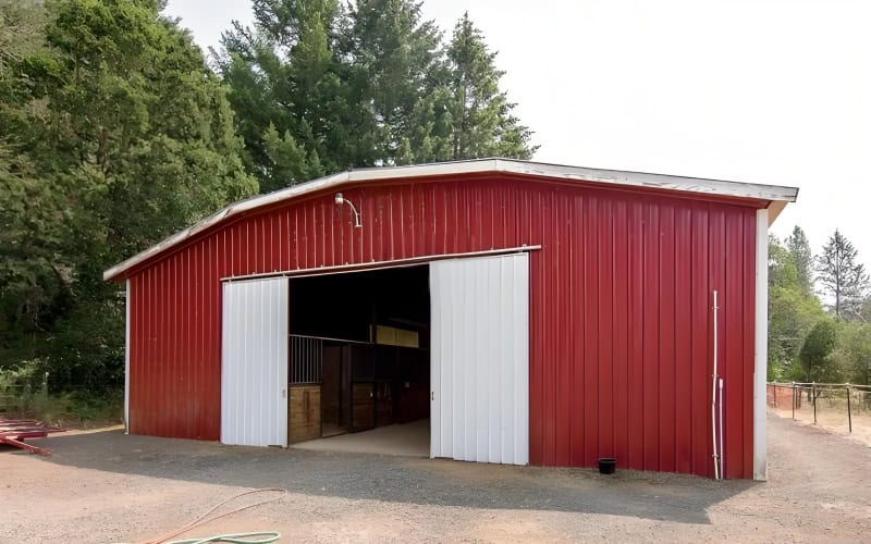 Another red pole barn with white sliding doors, surrounded by trees, and some items visible inside the open entrance.