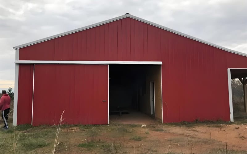 A red pole barn with a partially open sliding door, set on a grassy and dirt area under a cloudy sky.