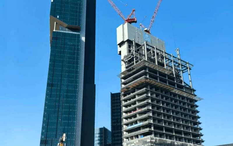 A view of a tall skyscraper under construction with a partially completed steel frame and cranes in the background.