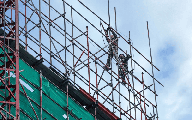 Workers assembling a scaffolding structure on a high-rise building with safety equipment visible.