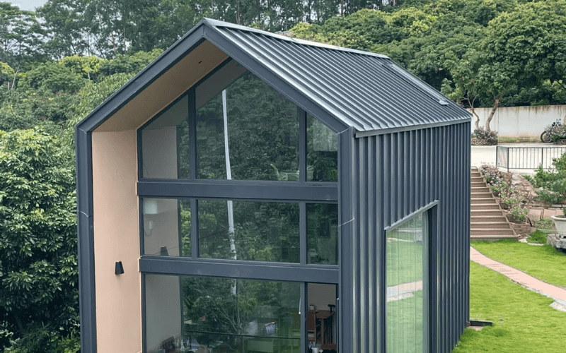 A modern steel-frame building with large windows and a sloped metal roof, surrounded by green landscaping.