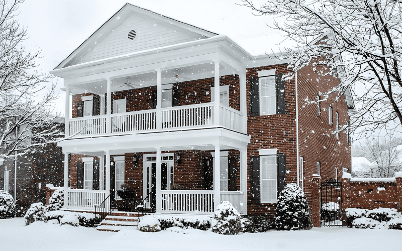 A classic brick house covered in snow, with white pillars and a snow-covered yard. The scene is peaceful and wintery with snowflakes falling from the sky.