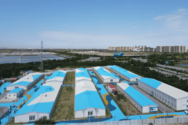 An aerial view of a factory transformer facility with blue and white roofs, surrounded by greenery and nearby infrastructure, under a clear blue sky.